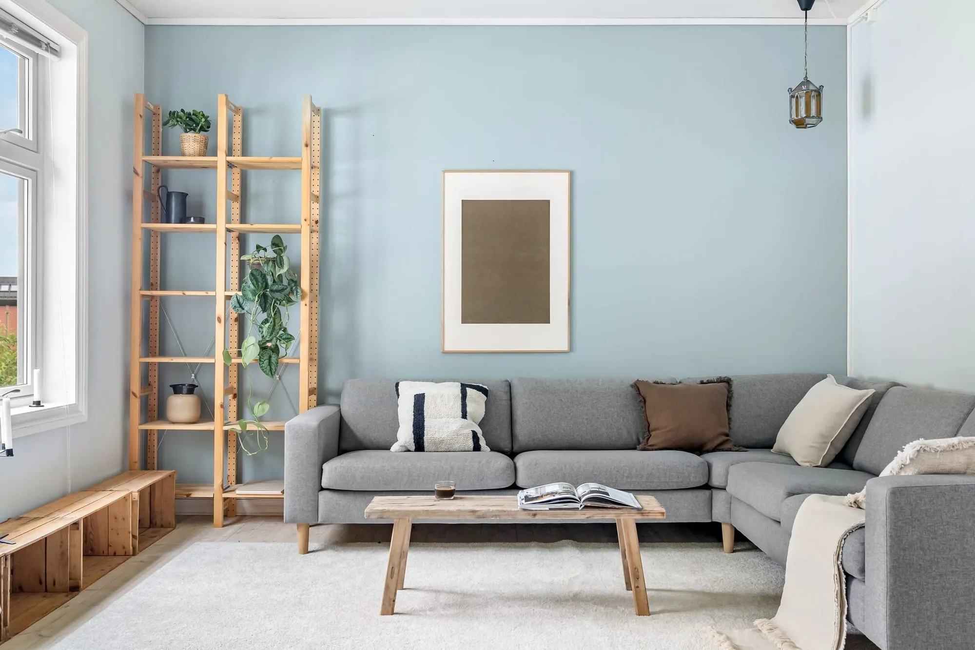 Helles Wohnzimmer mit hellblauer Wand, grauem Sofa und Holzregal am Fenster